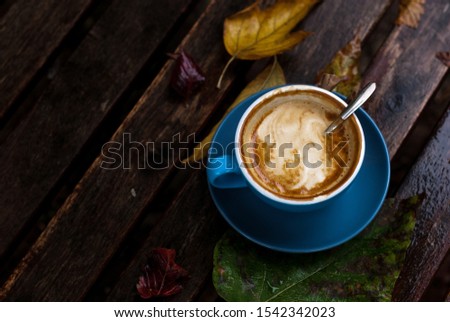 Blue coffee cup filled with coffee and cream on top, on a autumn rainy background with wooden table and leaves