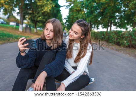Two girls schoolgirls teenagers 12-15 years old, autumn day summer city park, smartphone, selfie photo, video call, sweaters everyday clothes. Emotions joy fun, happy smiling sitting skateboard