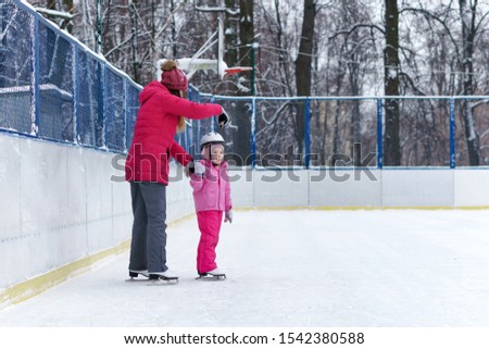 Mom teaches her little daughter to skate on the rink on a winter day. Weekends activities outdoor in cold weather.