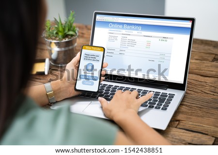 Woman Using Mobile Phone App To Authenthificate Bank Transfer On Laptop Royalty-Free Stock Photo #1542438851