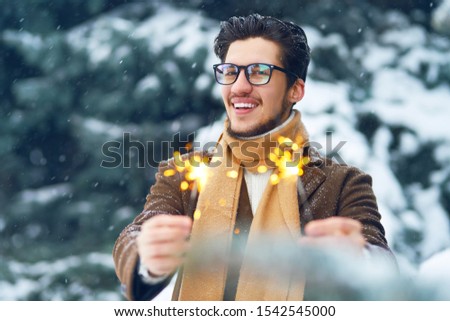 Young man in a coat on the street with sparklers. Smiling young man having fun outdoors. Man enjoy a winter. Dressed in a coat, sweater and scarf. Winter concept. Snowfall.