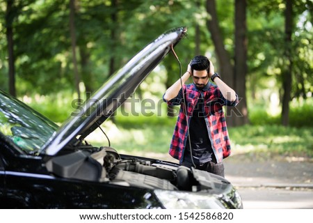 Closeup portrait, young man having trouble with his broken auto, opening hood trying to fix engine, isolated green trees outside background. Car won't start, dead battery Royalty-Free Stock Photo #1542586820