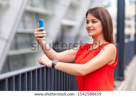 I am beautiful. Half turned portrait of attractive brunette woman in stylish red shirt doing selfie using smartphone, confident in her beauty looking stunning, taking picture for blog, outdoor shot