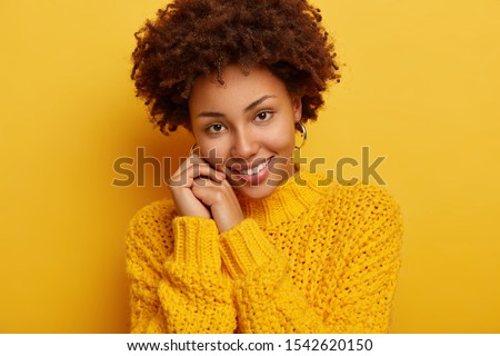 Headshot of adorable romantic woman has pleasant smile, tilts head on hands, has tender look, has dark curly hair, wears cozy winter sweater, isolated over yellow background. Happy emotions.