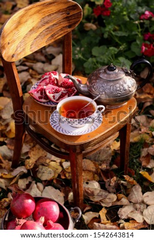 Fresh cup of black tea, old aged metal silver-plate teapot, pattern roses, pomegranate on weathered wooden child chair on autumn leaves, natural background, composition in garden, outdoor and space