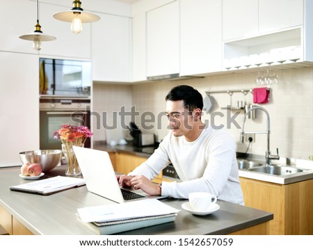 young asian man working from home sitting at kitchen counter using laptop computer