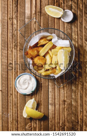 Classic british fast food fish and chips served in frying basket with lemons, white sauce, salt in ceramic bowls over wooden plank background. Flat lay, space