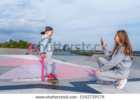 Woman mom takes pictures on phone, support care training, little boy 3-5 years old son stands on skateboard, trains in summer to ride city, autumn on sports ground, happy smiling casual clothes