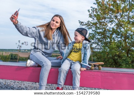 Woman mom takes pictures selfie phone little boy 3-5 years old son, sits resting, summer city autumn sports ground happy smiling casual clothes. Skateboard video call smartphone application Internet
