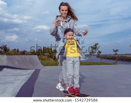 Happy family mother woman little boy son 3-5 years old, summer sports field city, playing skateboarding, emotions joy, fun relaxation weekend. Everyday clothes, parenting support care child