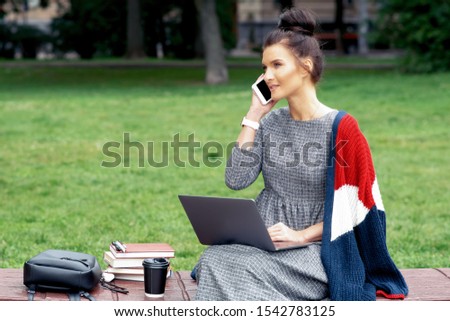 Smiling female student talks by smartphone at park. Student girl is talking by smartphone with laptop at the park.