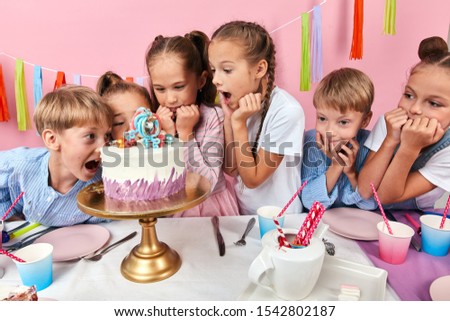 little girls and boys with wide open mouths loking at the cake with amazed expression. isolated pink background