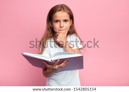 little clever girl touching her chin , going to read a book, kid being puzzled with the plot of story, hobby, lifestyle, free time, spare time. isolated pink background, studio shot