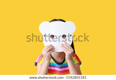 Little child girl holding blank white animal paper mask fronting her face isolated on yellow background. Idea and concept for kid dressed up playing animal face.