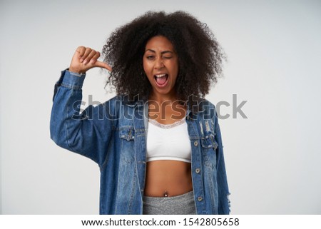 Studio shot of cheerful pretty young female with dark skin winking at camera with joyful broad smile, wearing white top and jeans coat, isolated over white background with raised hand