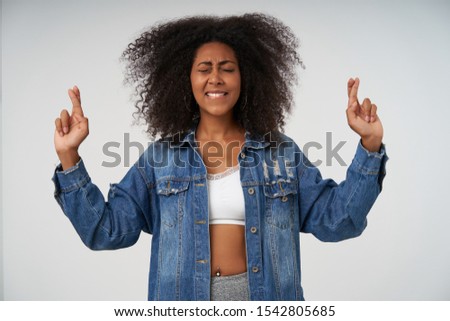 Indoor shot of positive young curly dark skinned lady raising hands with crossed fingers, making wish and keeping eyes closed, isolated over white background in casual clothes