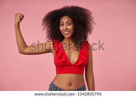 Positive pretty curly dark skinned female with belly button piercing looking at camera with wide cheerful smile, demonstrating her power in raised hand, posing over pink background