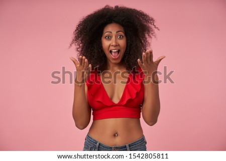 Overjoyed young curly woman with dark skin wearing casual clothes standing over pink background, raising palms happily and looking at camera with wide eyes and mouth opened