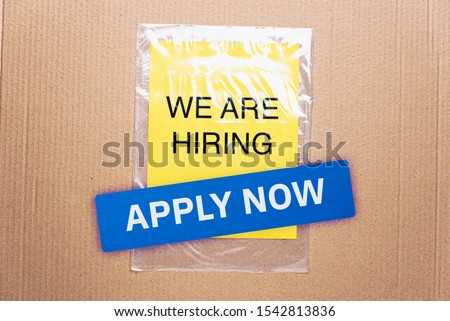 Apply now job button here hiring resources register online concept. We are hiring printed on yellow paper on transparent plastic bag with cardboard background.