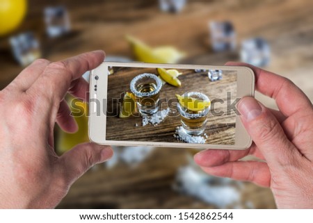 Man taking photo of Mexican traditional alcohol drink Tequila with lemons and salt and ice cube on wood table