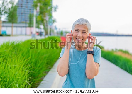 Portrait of smiling senior woman listening to music after running. Healthy mature woman wearing blue headband and sportswear adjusting earphones. Closeup face of retired woman jogging at park.