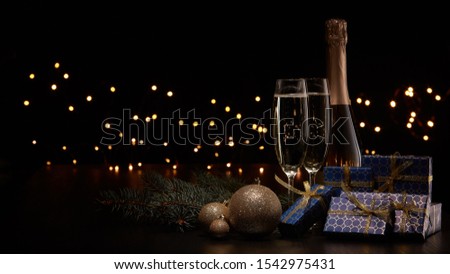 Still life with champagne bottle, two champagne flutes, gift boxes, fir branch, Christmas ornaments and LED lights garland on black background with copy space. Christmas and New Year.  Greeting card
