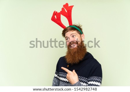 Redhead man with long beard dressed up for christmas holidays over isolated green background pointing to the side to present a product