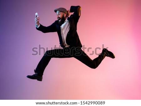 Full length portrait of happy jumping man wearing office clothes in neon light isolated on gradient background. Emotions, ad concept. Using smartphone, winning bet or sale, shopping, talking