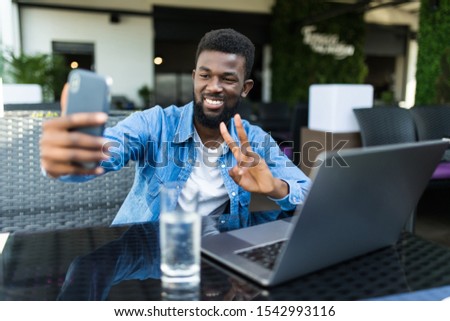 Young african man with phone take selfie make peace gesture while sitting with laptop