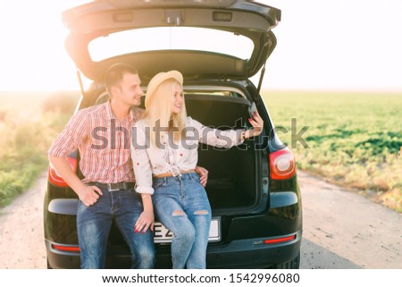 Young couple in sweaters sitting together in the car trunk and taking selfie photo on the roadside in the forest.