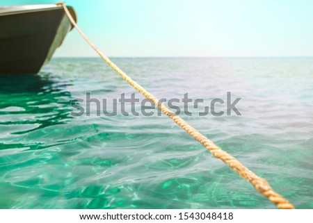 A thick clothesline is tied to an anchor that lies at the bottom of a clear blue sea, keeping the green boat on the surface in Sunny weather with clouds and glare, calm blue sea, small waves