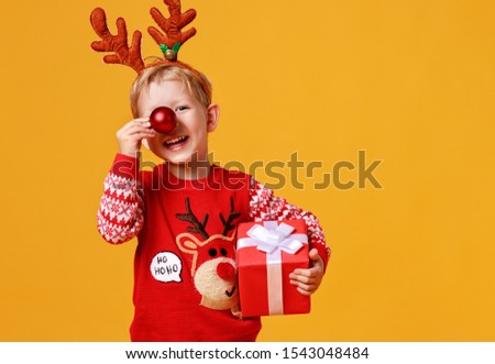happy funny child boy in red Christmas reindeer costume with gift on yellow colored background
