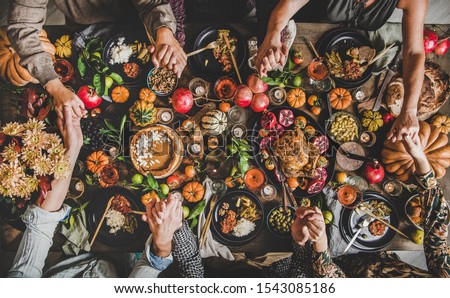 Family praying holding hands at Thanksgiving table. Flat-lay of feasting peoples hands over Friendsgiving table with Autumn food, candles, roasted turkey and pumpkin pie over wooden table, top view