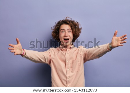 Emotive caucasian young man dressed in light coloured shirt with long slaves, stretching palms towards camera, happy to meet good friend, opens mouth with joyful feelings, shout loudly
