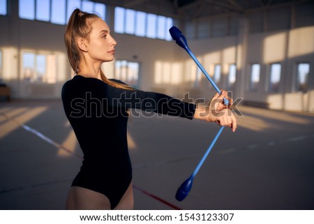 Close view of female gymnast playing with blue clubs in roomy studio, looking away, side shot, professional sport concept