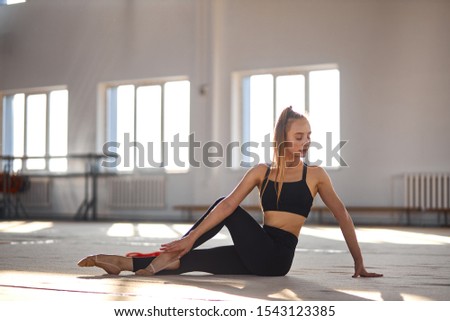 Young pretty rhythmic gymnast training and stretching on the floor, posing against background of gymnastics school