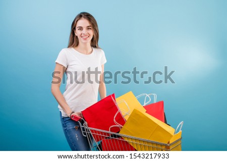 brunette woman with shopping cart full of colorful paper bags isolated over blue