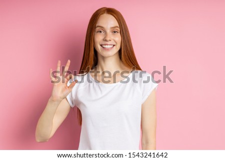 Photo of cheerful young redhead woman wearing t shirt making okay gesture, demonstrating her agreement, smiling with perfect white teeth isolated over pink background. Body language concept.