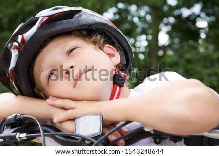 Small Caucasian boy cyclist in protective helmet put his head on the handlebar of the bike posing for the camera. a Boy rests during cycling in park on summer day. Weekend activity.