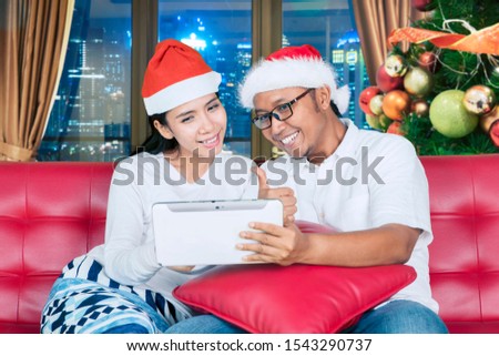Young couple wearing wearing Santa hat while showing thumb up and using a digital tablet on the couch. Christmas online shopping concept
