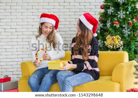 Smiling beautiful two women with santa hat opening gift box sitting on yellow sofa in white brick background - Asian young pepole in christmas party concept