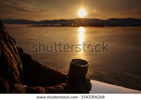 Thermo mug with tea in the hands of man against the sunset sky and sea, close-up