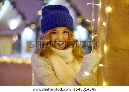 Smiling woman with garlands and holiday lights on festive Christmas fair. Lady wearing classic stylish winter knitted clothes.