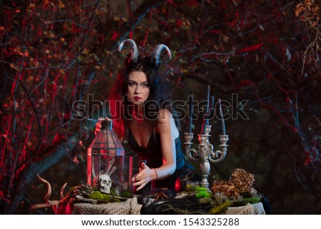 Beautiful witch with horns on her head is being prepared to make the witchcraft over the forest background. Halloween image.