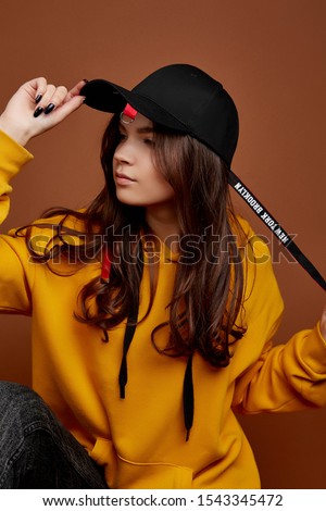 Half-turn shot of European lady with long brown hair in a yellow hoodie, black jeans and a black baseball cap with a red ribbon and band with lettering "New York Brookllyn".