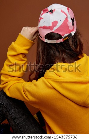 Back view shot of a brunette lady with long brown hair in a yellow hoodie and black jeans and colorful baseball cap with a camouflage print. The girl is posing on the brown background. 
