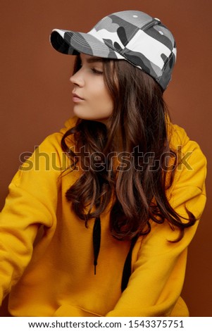 Profile photo of European lady with long brown hair in a yellow hoodie and a colorful baseball cap with camouflage print. The girl is posing on the brown background.