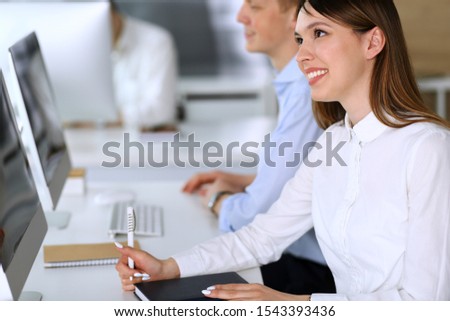 Cheerful smiling asian businesswoman headshot at work in modern office. Casual dressed female entrepreneur using pc computer while sitting with diverse colleagues at the background. Multi ethnic