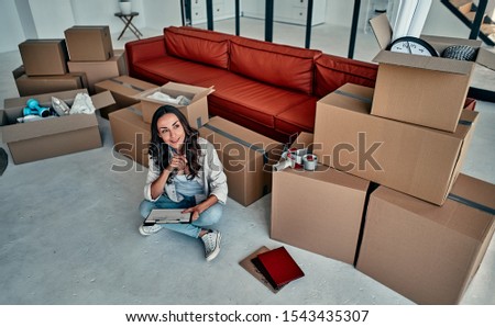 A young woman in the living room at home unpacking boxes with things. A woman with a folder is thinking about a new design. Moving, buying a house, apartment concept.
