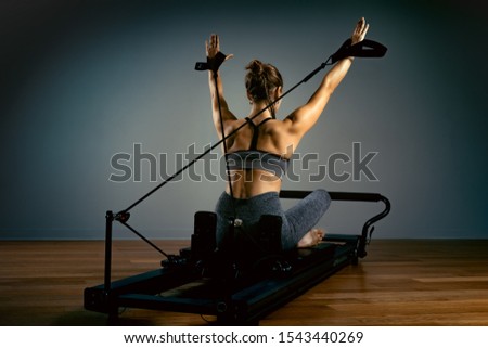 Young girl doing pilates exercises with a reformer bed. Beautiful slim fitness trainer on reformer gray background, low key, art light. Fitness concept Royalty-Free Stock Photo #1543440269